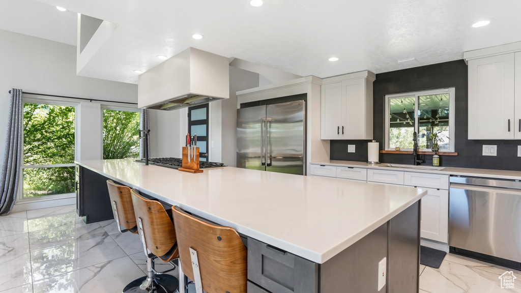 Kitchen with appliances with stainless steel finishes, light marble finish flooring, custom exhaust hood, a spacious island, and white cabinetry
