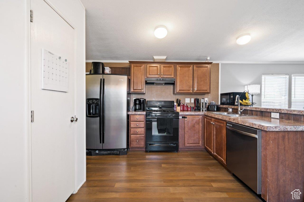 Kitchen with appliances with stainless steel finishes, brown cabinetry, dark wood finished floors, a peninsula, and under cabinet range hood