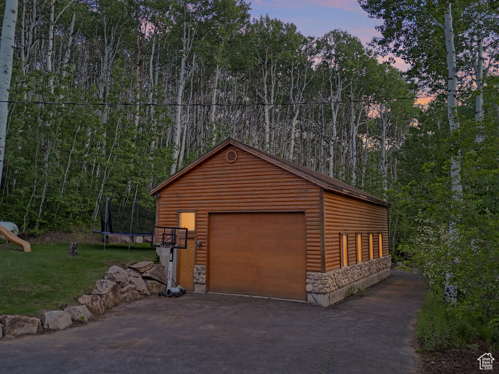 Garage at dusk featuring a trampoline, a garage, a lawn, asphalt driveway, and a forest view