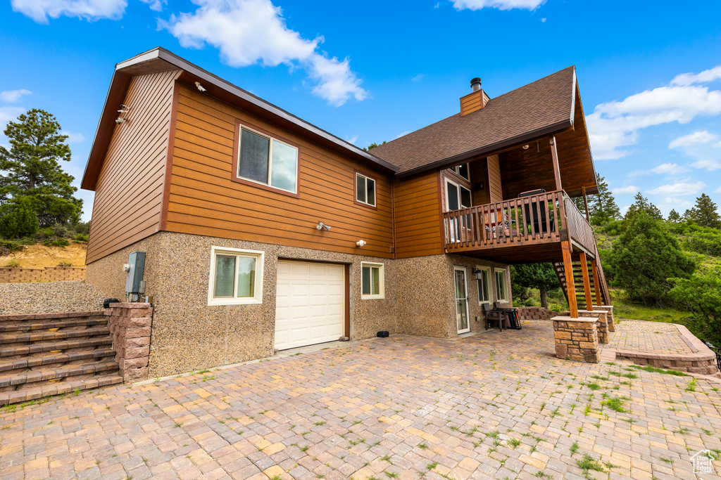 Rear view of house featuring an attached garage, stairs, a chimney, a wooden deck, and a patio