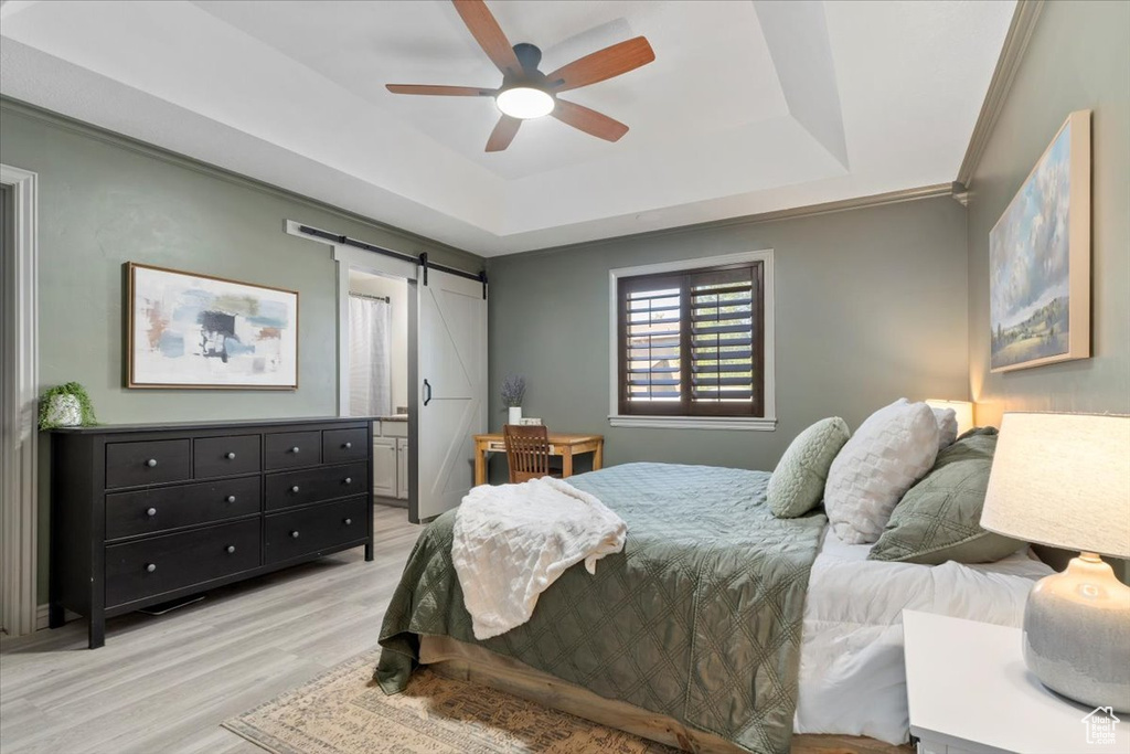 Bedroom with a barn door, a raised ceiling, light wood-style flooring, a ceiling fan, and crown molding