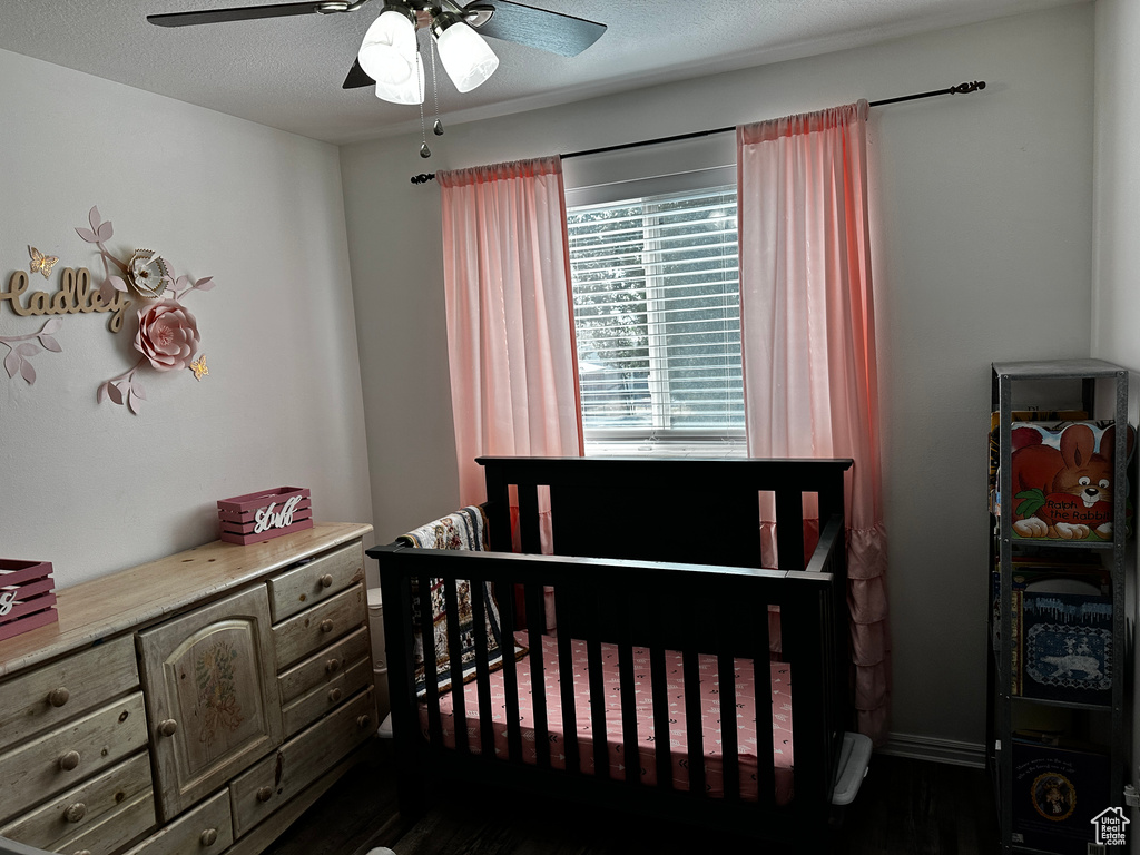 Bedroom with dark wood-style floors, a nursery area, a ceiling fan, and a textured ceiling