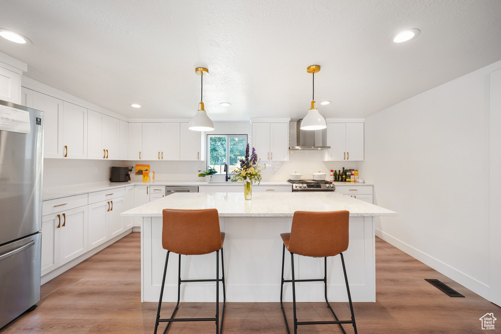 Kitchen featuring appliances with stainless steel finishes, wall chimney range hood, a center island, a breakfast bar area, and white cabinets