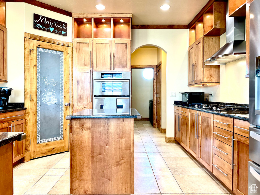 Kitchen with arched walkways, wall chimney range hood, stainless steel double oven, a kitchen island, and light tile patterned floors