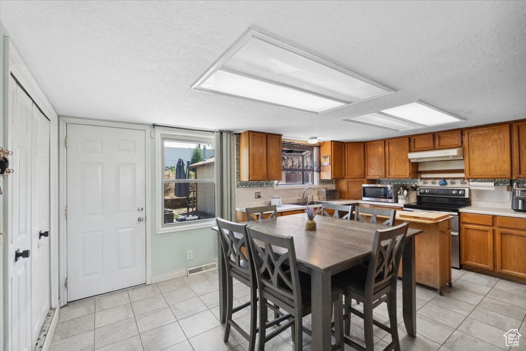 Kitchen featuring appliances with stainless steel finishes, under cabinet range hood, light tile patterned flooring, brown cabinetry, and a textured ceiling