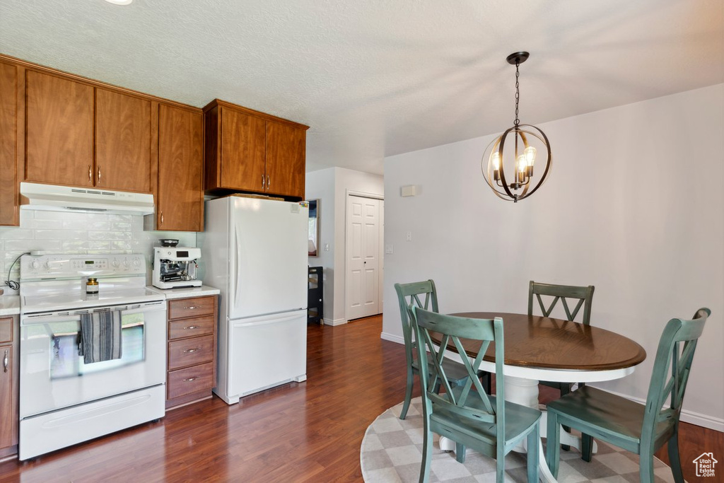 Kitchen with under cabinet range hood, white appliances, brown cabinets, a chandelier, and dark wood finished floors