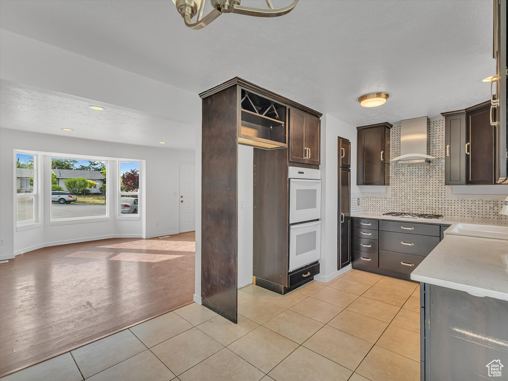 Kitchen featuring wall chimney exhaust hood, dark brown cabinets, white appliances, light tile patterned floors, and backsplash