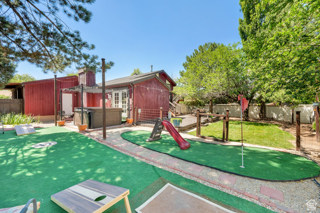 View of jungle gym featuring an area to practice putting, a fenced backyard, and a hot tub
