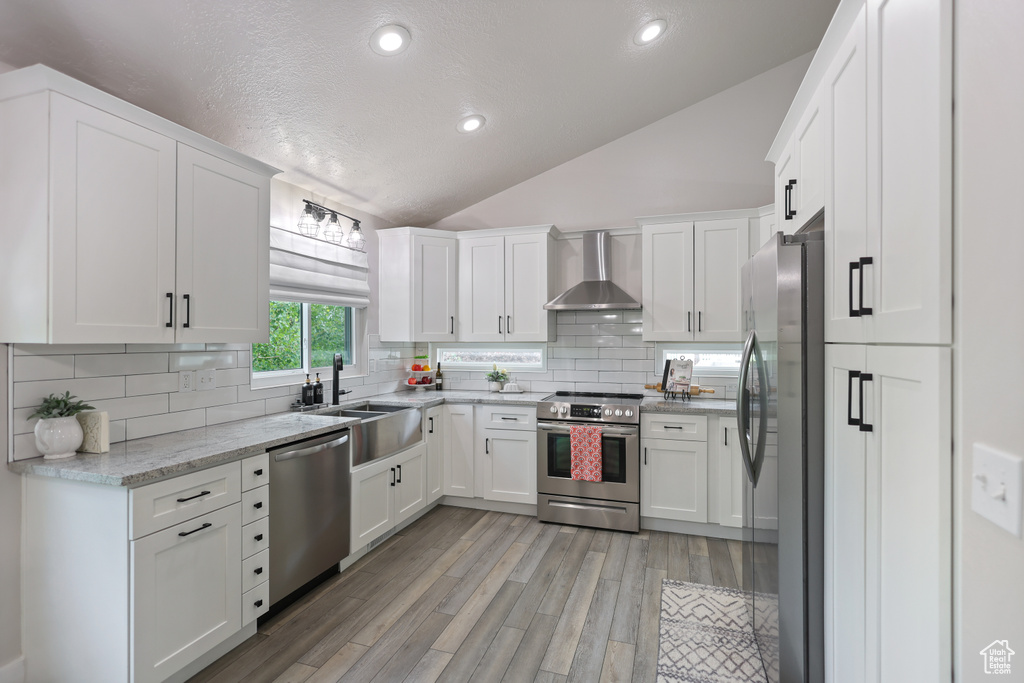 Kitchen featuring appliances with stainless steel finishes, wall chimney exhaust hood, vaulted ceiling, a sink, and white cabinets