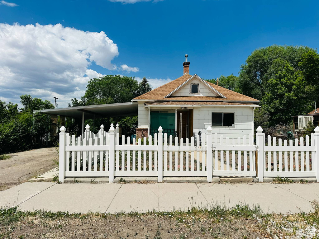 Bungalow-style home with a fenced front yard, a shingled roof, a chimney, and a carport