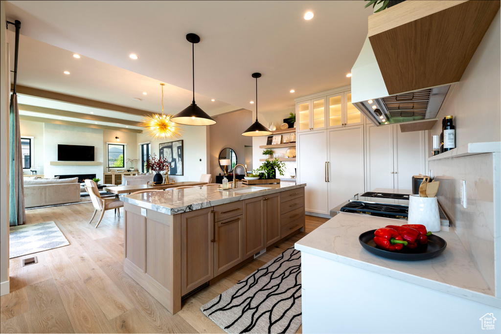 Kitchen featuring open shelves, wall chimney range hood, light wood-style floors, an island with sink, and light stone counters