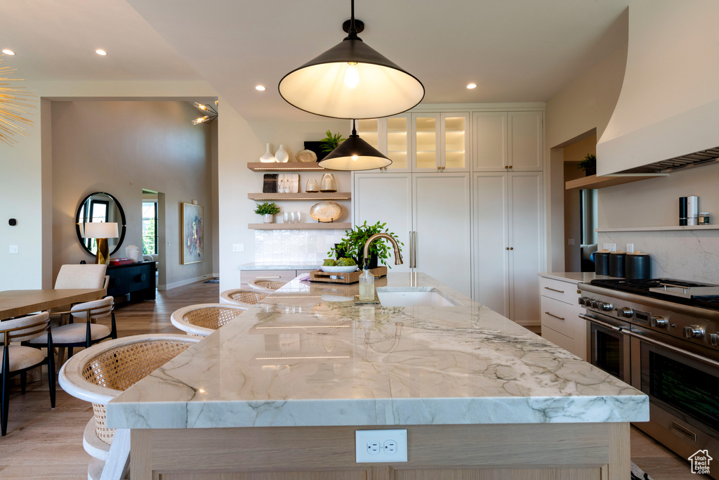 Kitchen featuring range with two ovens, premium range hood, open shelves, white cabinets, and a center island with sink