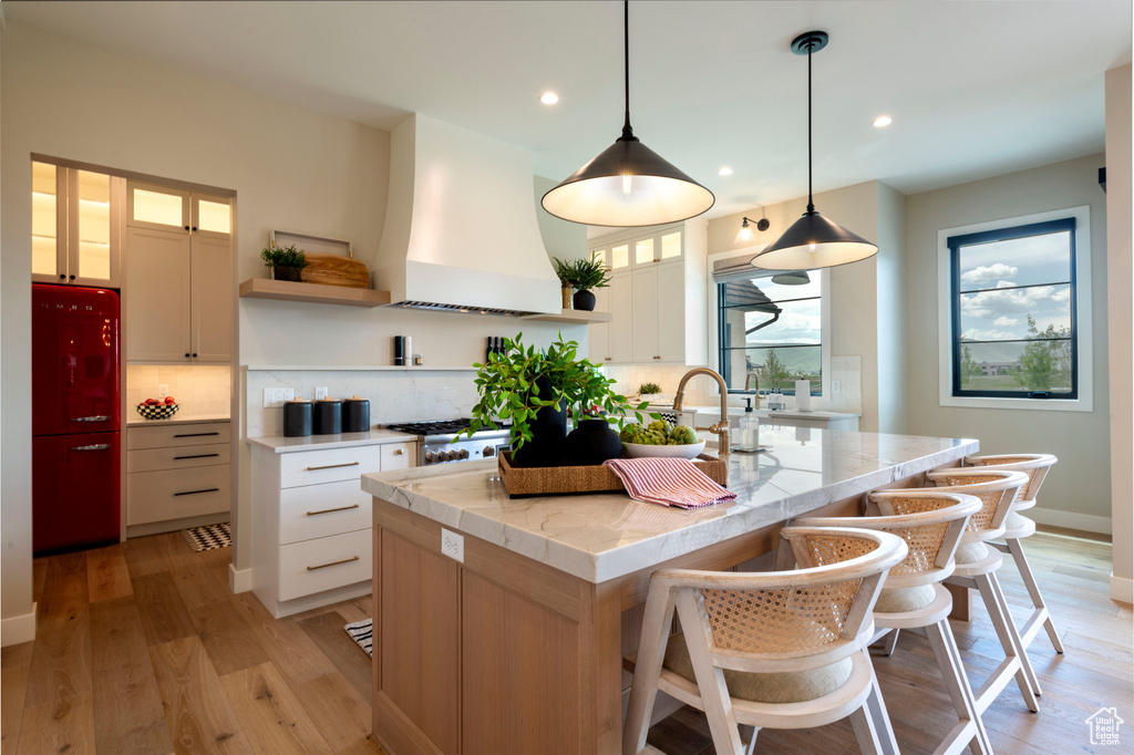 Kitchen with open shelves, premium range hood, a kitchen island with sink, backsplash, and light wood-type flooring