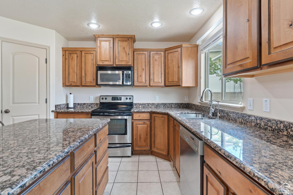Kitchen featuring appliances with stainless steel finishes, dark stone countertops, brown cabinetry, a textured ceiling, and light tile patterned floors