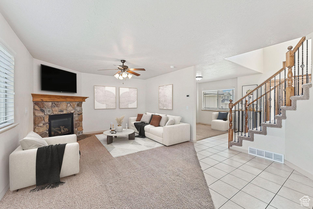 Living area featuring a ceiling fan, a stone fireplace, light carpet, stairs, and a textured ceiling