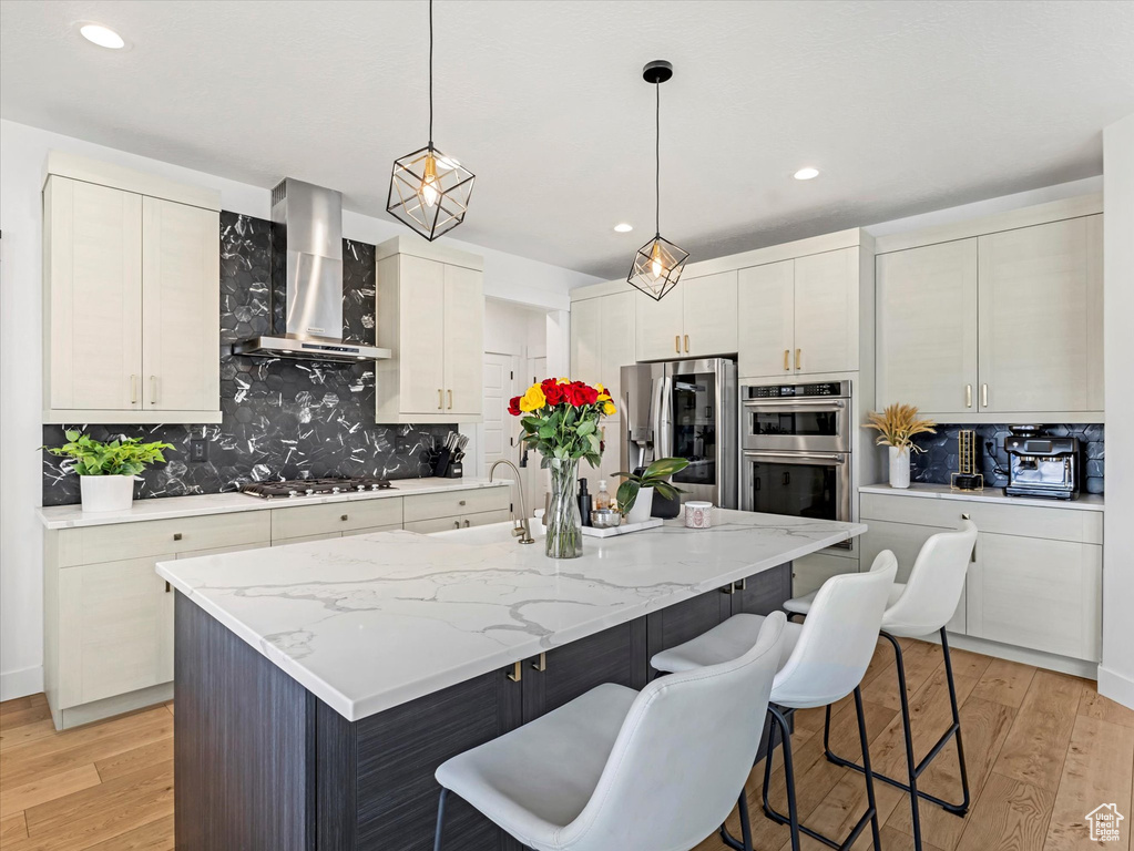 Kitchen featuring wall chimney range hood, stainless steel appliances, a breakfast bar area, light wood finished floors, and decorative backsplash