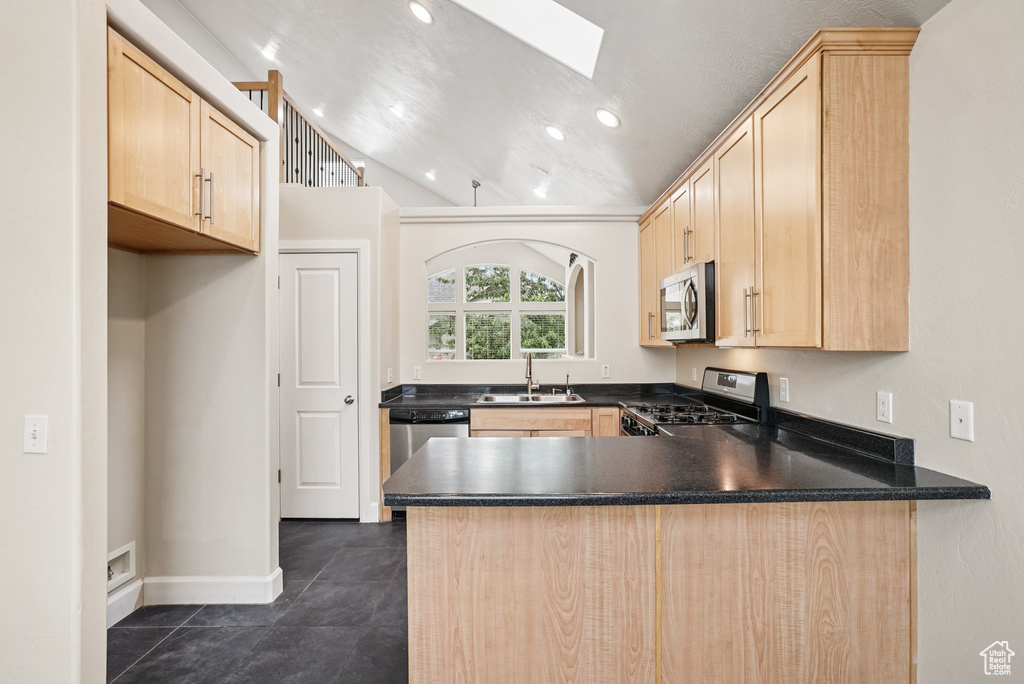 Kitchen featuring appliances with stainless steel finishes, light brown cabinetry, a peninsula, dark countertops, and a skylight