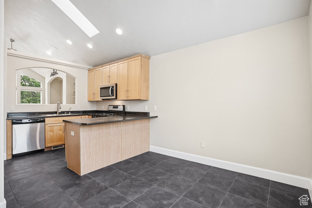 Kitchen featuring stainless steel appliances, light brown cabinetry, a skylight, a peninsula, and dark countertops