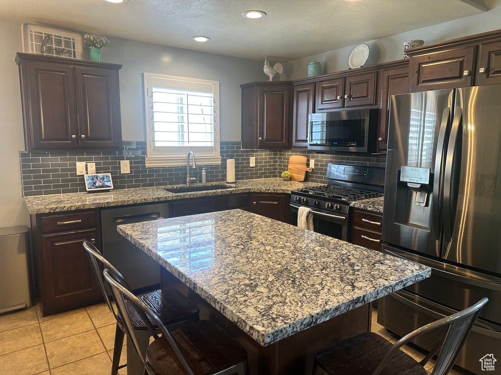 Kitchen with appliances with stainless steel finishes, a breakfast bar area, dark brown cabinetry, decorative backsplash, and light stone countertops
