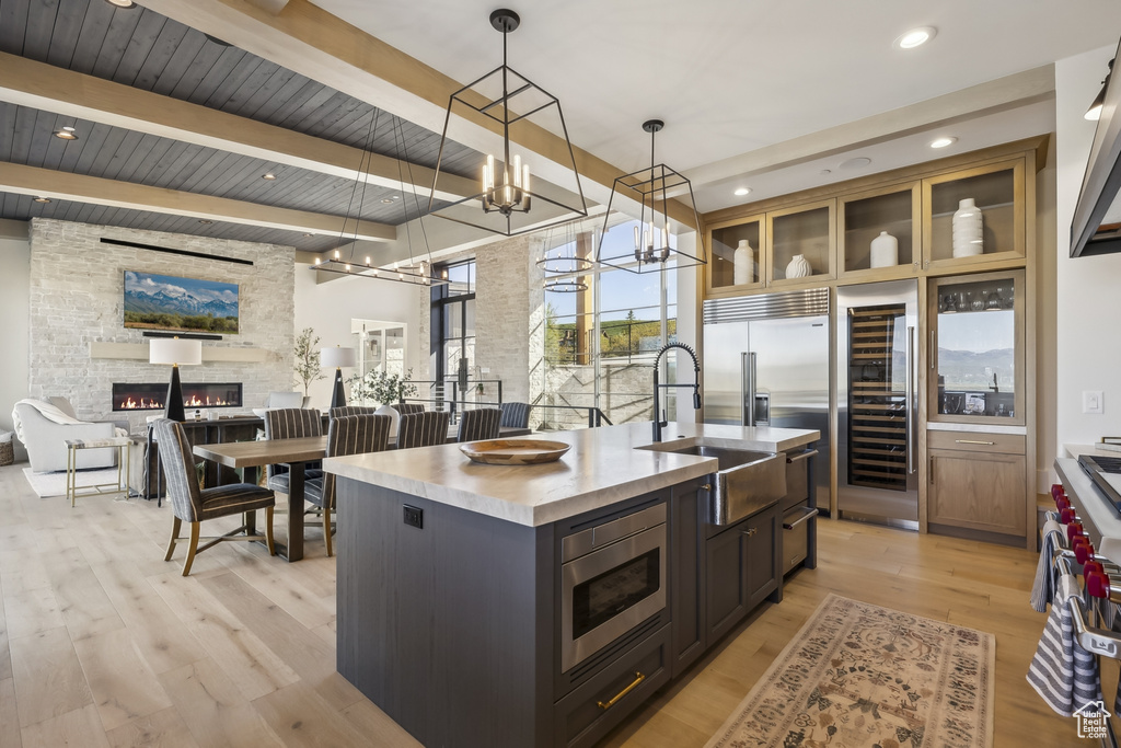 Kitchen featuring high end appliances, a stone fireplace, a center island with sink, light wood-style floors, and decorative light fixtures