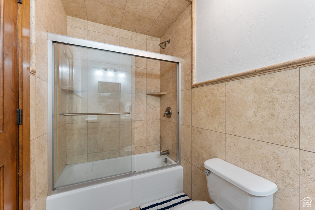 Bathroom with tile walls, bath / shower combo with glass door, and toilet