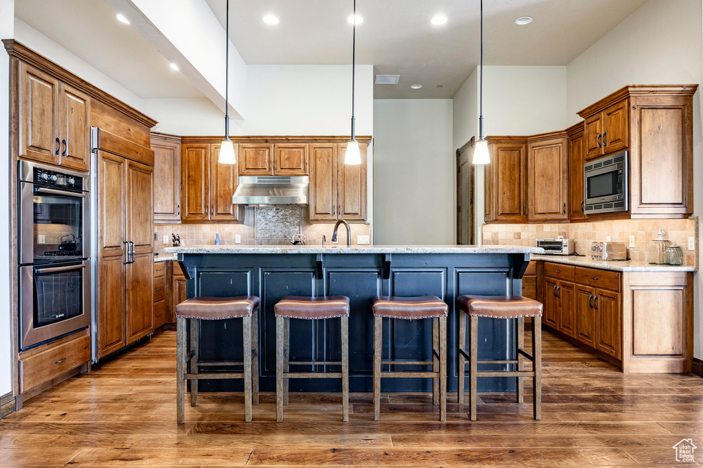 Kitchen featuring under cabinet range hood, built in appliances, an island with sink, brown cabinetry, and tasteful backsplash