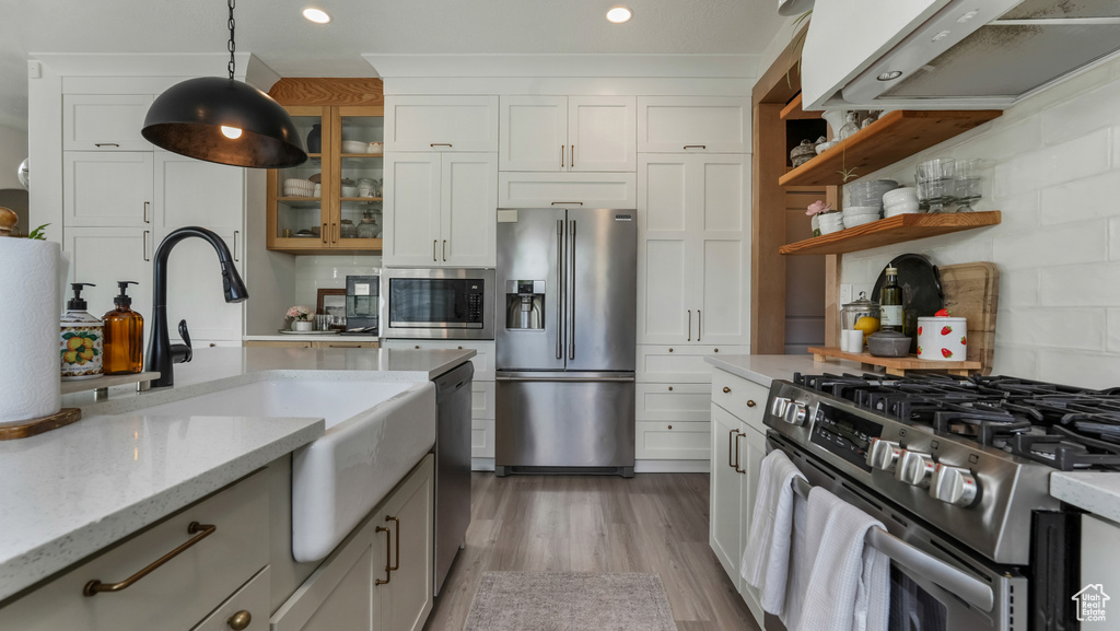 Kitchen featuring appliances with stainless steel finishes, ventilation hood, glass insert cabinets, light wood-style floors, and decorative light fixtures