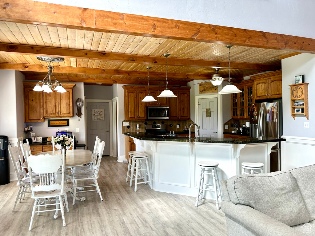 Kitchen with wooden ceiling with exposed beams, stainless steel appliances, a kitchen breakfast bar, light wood finished floors, and dark countertops