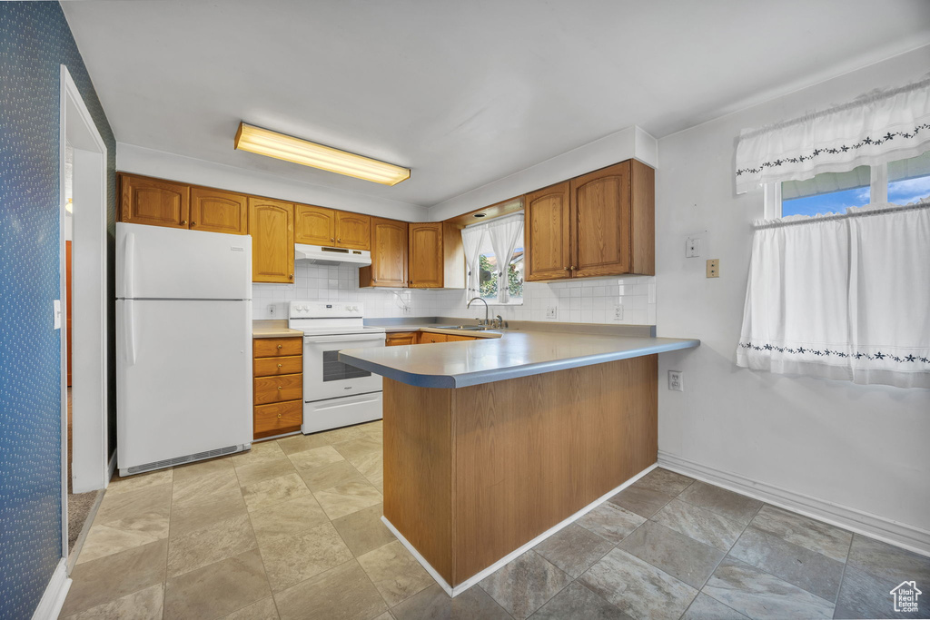 Kitchen with white appliances, under cabinet range hood, a peninsula, brown cabinets, and light countertops