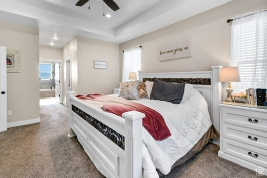 Bedroom with dark colored carpet, multiple windows, recessed lighting, ensuite bathroom, and a tray ceiling