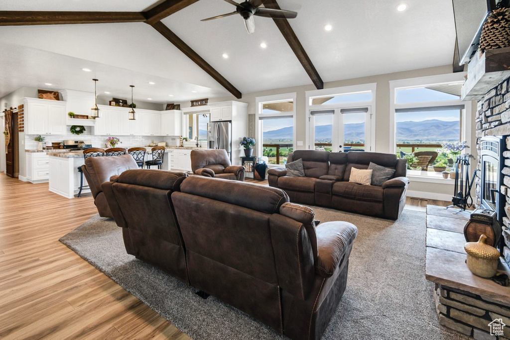 Living room featuring a ceiling fan, light wood finished floors, a stone fireplace, beamed ceiling, and recessed lighting