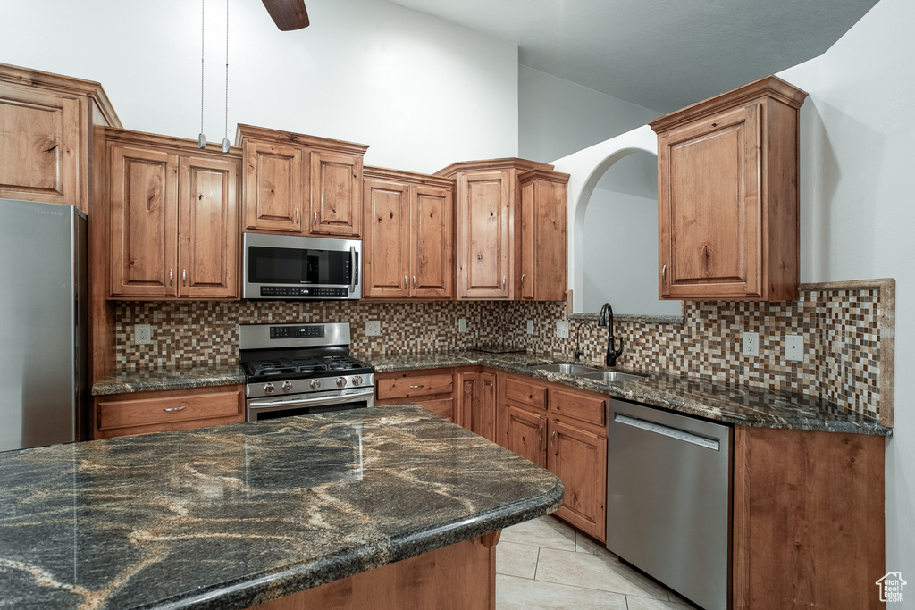 Kitchen with appliances with stainless steel finishes, brown cabinetry, decorative backsplash, dark stone countertops, and light tile patterned flooring