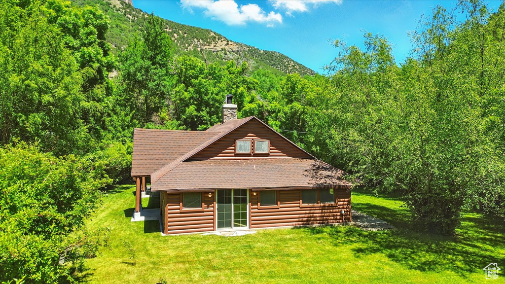 Back of house with a chimney, a forest view, roof with shingles, and a lawn
