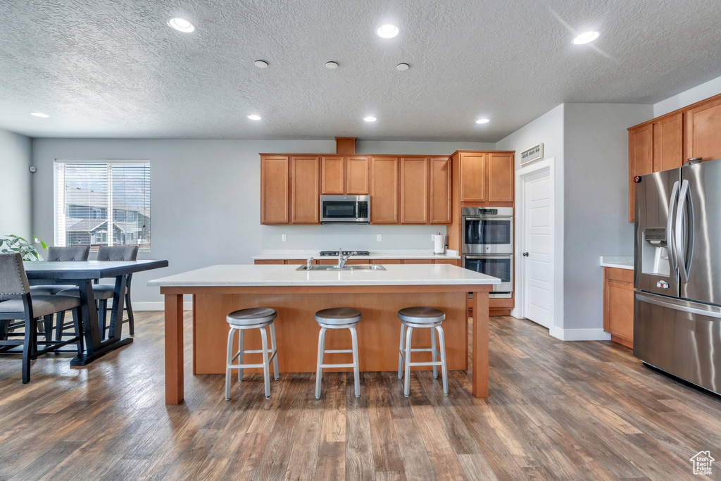 Kitchen featuring appliances with stainless steel finishes, a textured ceiling, an island with sink, light countertops, and dark wood-style floors