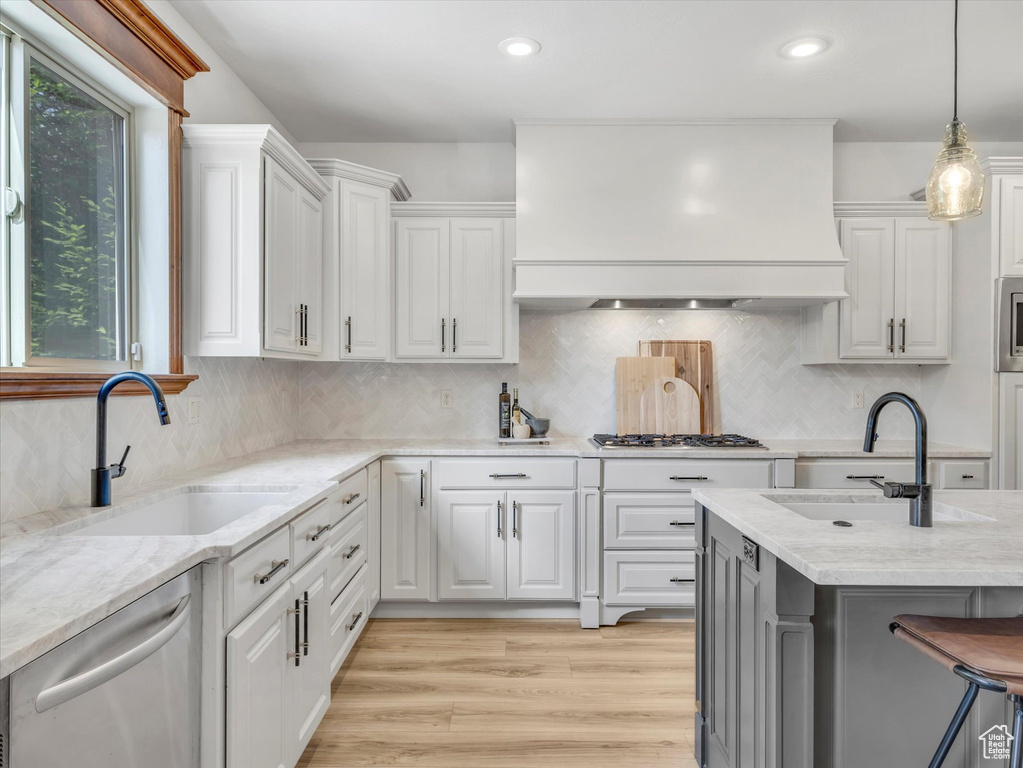 Kitchen with custom range hood, stainless steel appliances, white cabinetry, hanging light fixtures, and light wood-style floors