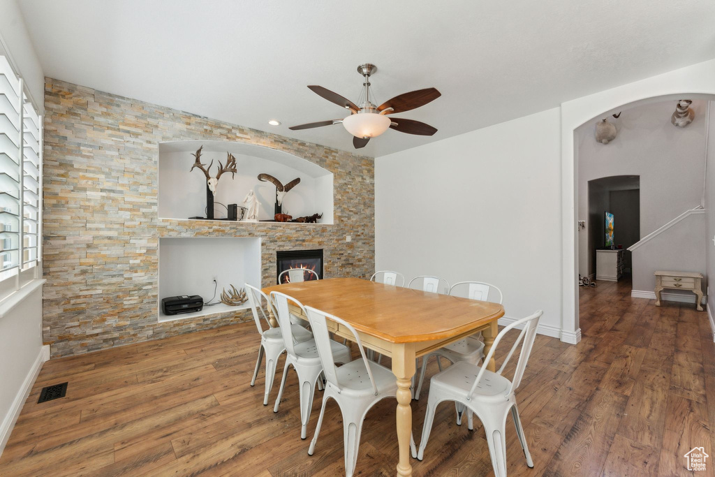 Dining area with ceiling fan, a stone fireplace, arched walkways, wood finished floors, and baseboards