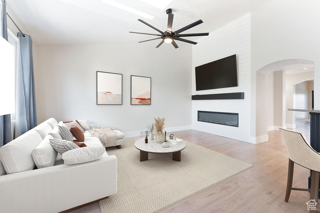 Living room with ceiling fan, arched walkways, light wood-type flooring, a large fireplace, and high vaulted ceiling