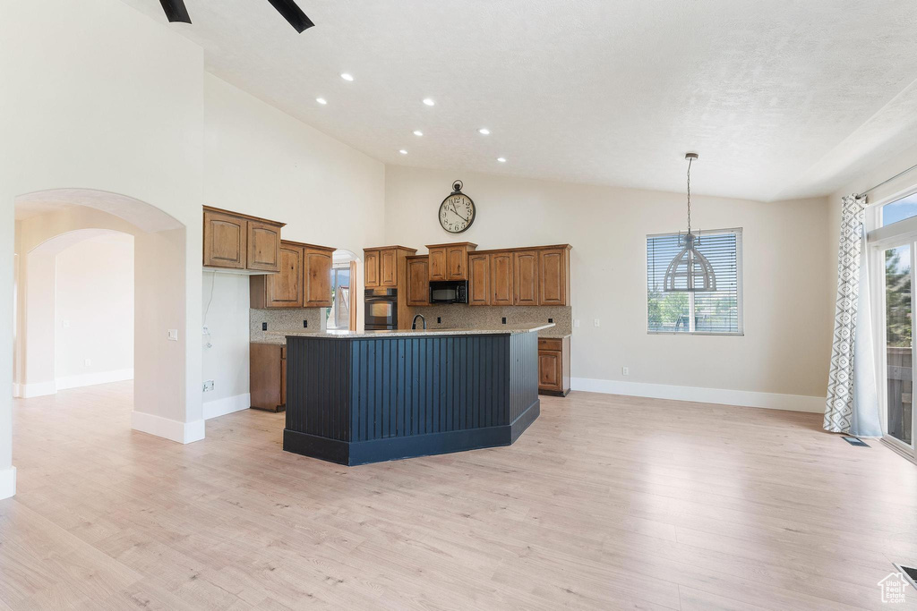 Kitchen with black appliances, brown cabinetry, a center island with sink, arched walkways, and light wood-style flooring