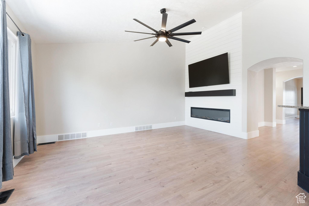 Unfurnished living room with a ceiling fan, arched walkways, light wood finished floors, and a large fireplace