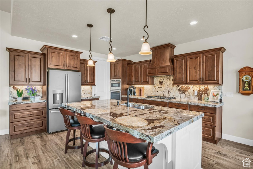 Kitchen with stainless steel appliances, custom range hood, light wood finished floors, a kitchen island with sink, and decorative backsplash
