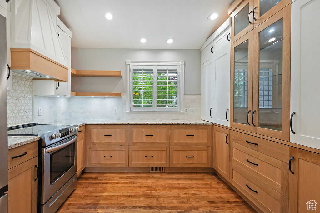 Kitchen featuring electric stove, custom exhaust hood, light stone counters, light wood finished floors, and glass insert cabinets