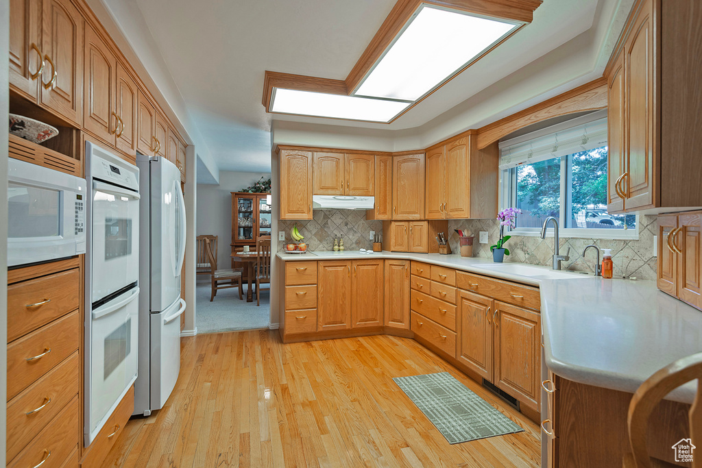 Kitchen with white appliances, under cabinet range hood, light wood-type flooring, light countertops, and decorative backsplash