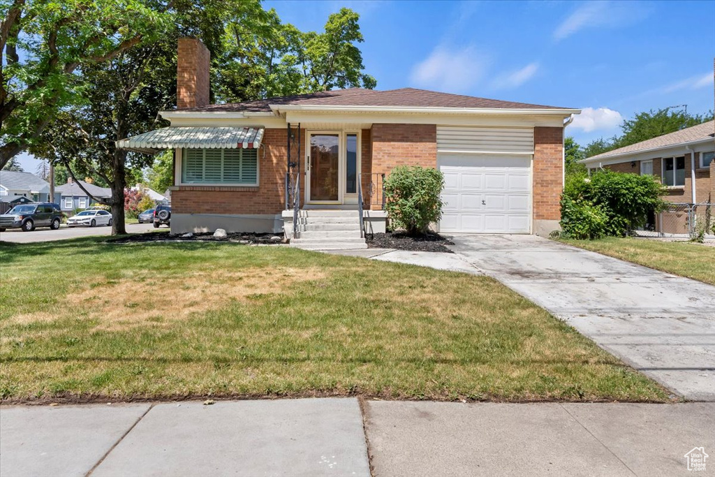 Single story home featuring an attached garage, a chimney, a front lawn, driveway, and brick siding