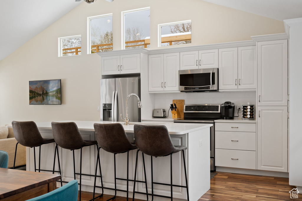 Kitchen featuring appliances with stainless steel finishes, a breakfast bar, a center island with sink, white cabinets, and high vaulted ceiling