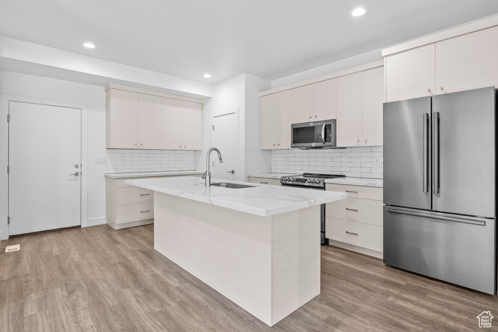 Kitchen with appliances with stainless steel finishes, decorative backsplash, light stone counters, light wood-style floors, and a center island with sink