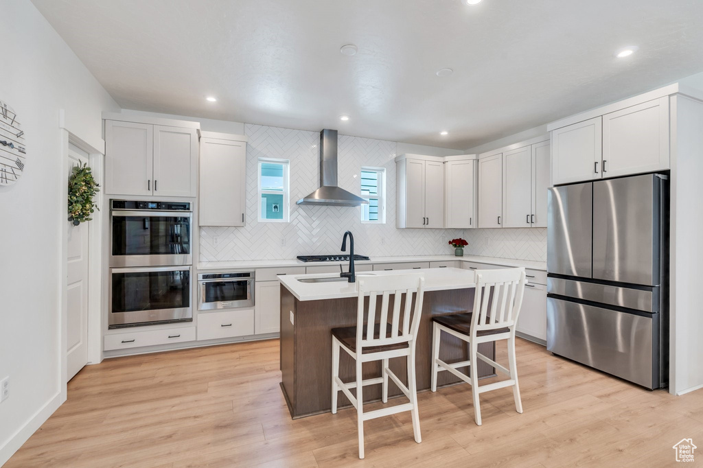 Kitchen with stainless steel appliances, wall chimney range hood, decorative backsplash, a center island with sink, and light countertops
