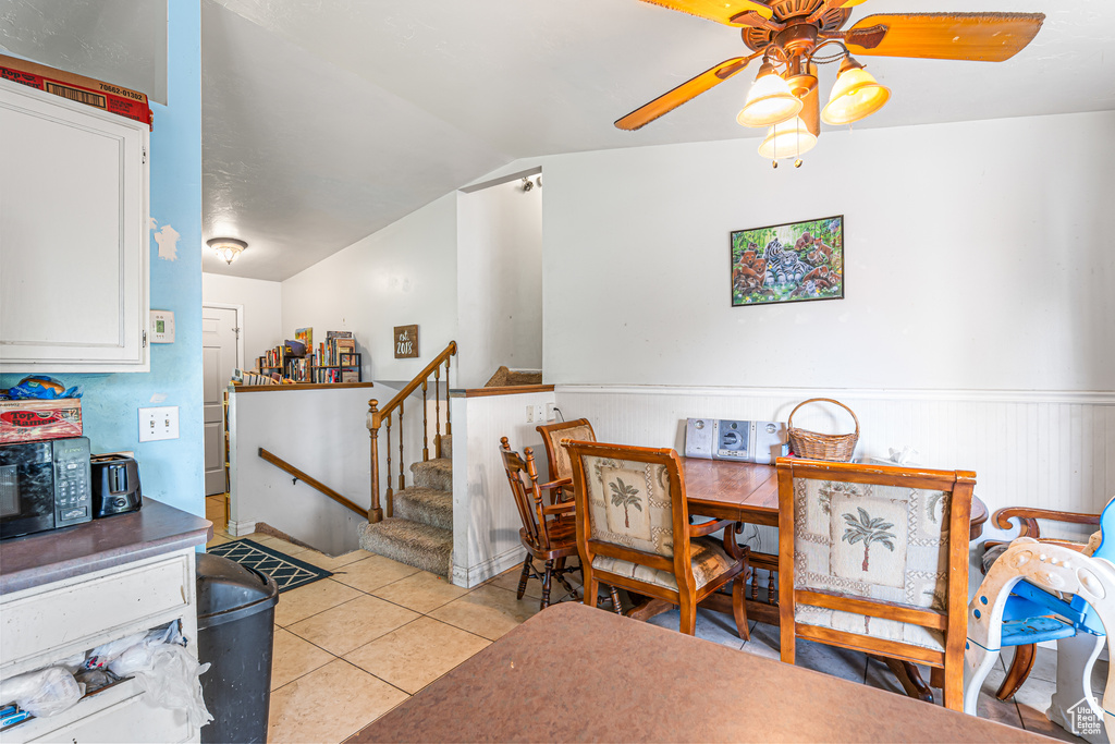 Dining space with a ceiling fan, wainscoting, light tile patterned floors, stairway, and vaulted ceiling