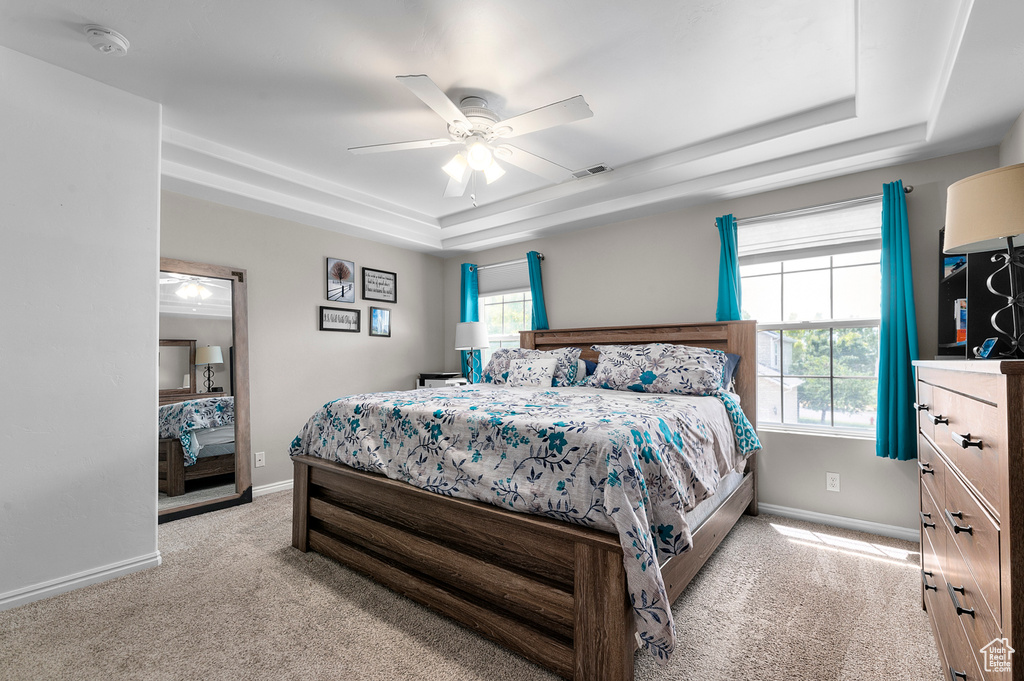 Bedroom with a tray ceiling, light colored carpet, multiple windows, and a ceiling fan
