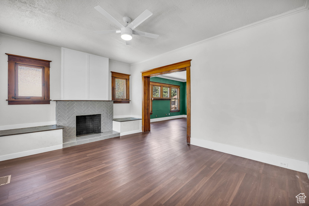 Unfurnished living room with dark wood-style floors, a tiled fireplace, ornamental molding, ceiling fan, and a textured ceiling