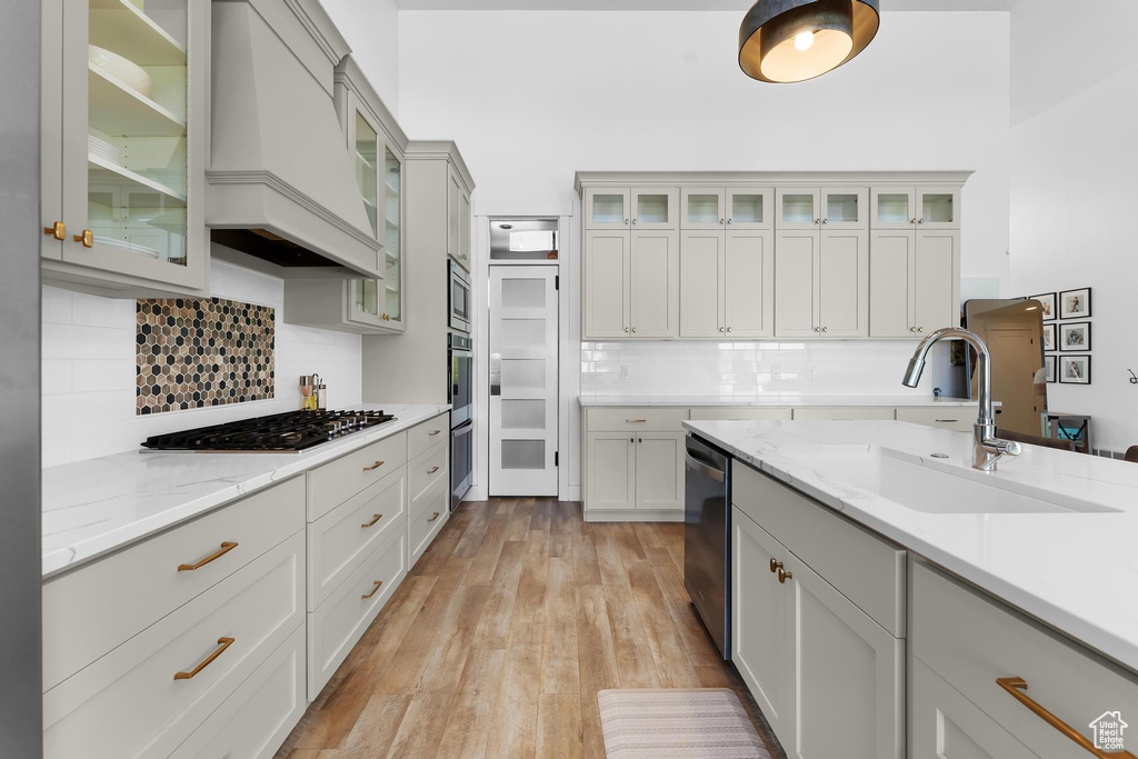 Kitchen featuring appliances with stainless steel finishes, custom exhaust hood, decorative backsplash, light wood-type flooring, and light stone countertops